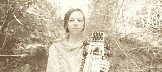 Jolanda Moyle holding a sarangi in front of a wooded background.