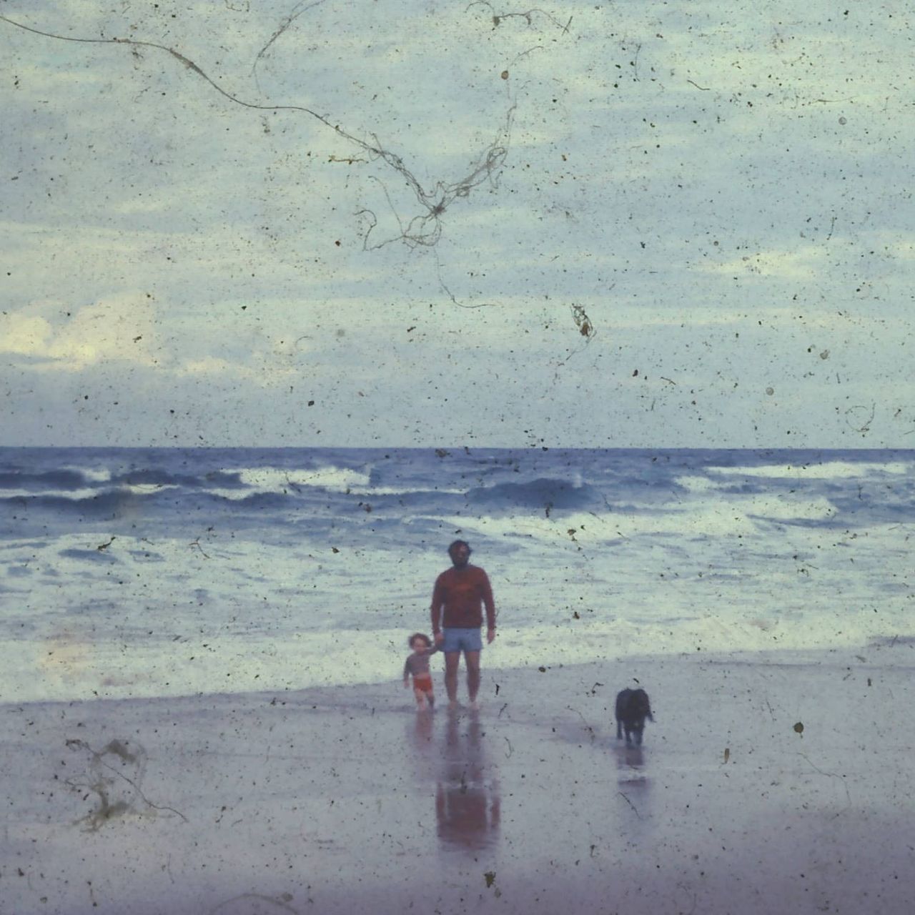 An old and degraded photograph of a boy, a man and a dog on a beach.